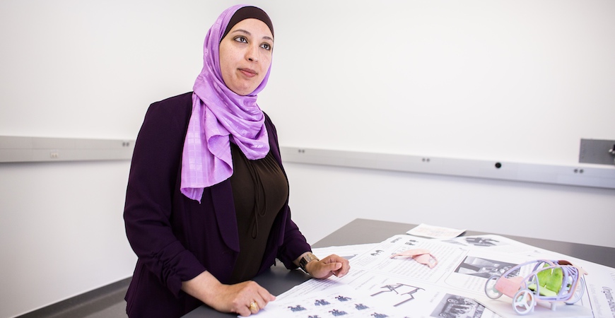 A woman in pink scarf and dark jacket stands in front of a table covered with posters and car prototypes.